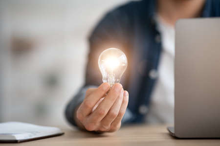 Unrecognizable man holding illuminated light bulb, sitting in officeの写真素材