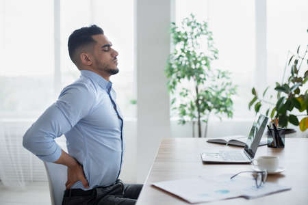 Young Arab businessman having acute back pain, massaging aching muscles, sitting at desk in modern office, copy spaceの写真素材