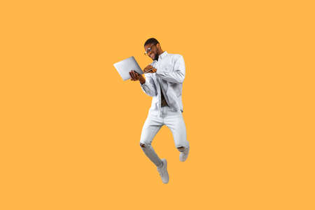 Happy african american man jumping up with laptop computer, having fun on yellow studio background