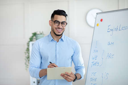 Positive male English teacher explaining foreign language rules near blackboard, taking notes in clipboard at officeの写真素材