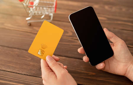 Online shopping concept. Woman holding credit card and smartphone with blank screen at wooden table...