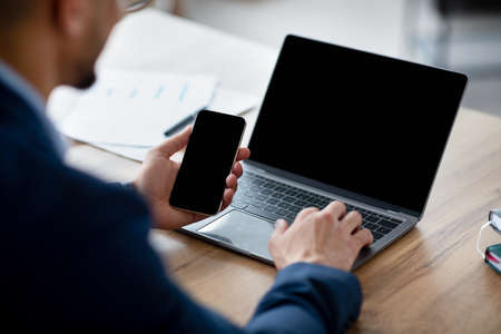 Cropped view of Arab businessman using laptop computer and smartphone with mockup for app or website...