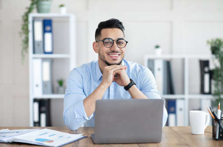 Portrait of professional young Arab guy sitting at his desk with laptop pc in modern office