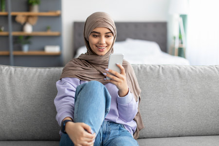 Smiling muslim woman using mobile phone sitting on couch