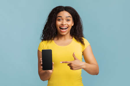 Joyful black lady pointing at smartphone with blank screen on blue studio background, mockup for...