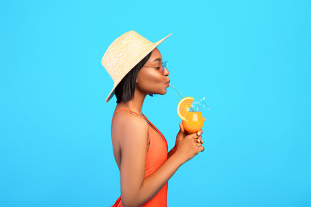 Side view of graceful black woman in swimsuit drinking cool refreshing tropical cocktail on blue studio backgroundの写真素材