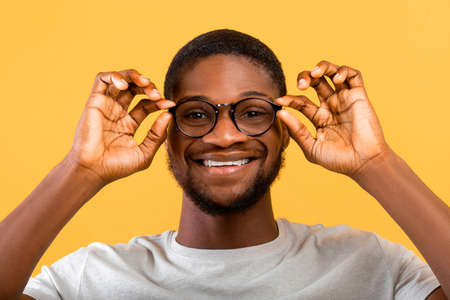 Positive african american guy wearing eyeglasses, looking and smiling to camera over yellow studio backgroundの写真素材