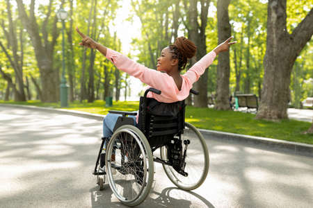 Handicapped black woman in wheelchair on walk at city park, showing victory gesture, feeling positive and motivatedの写真素材