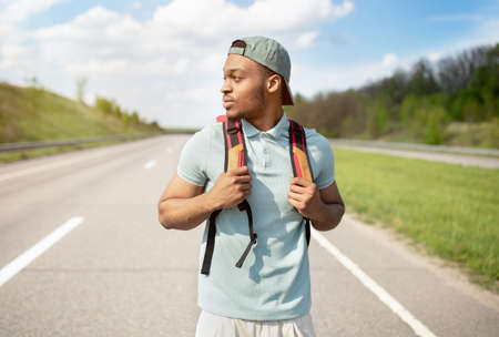 Summertime adventure. Handsome young black man with rucksack walking along asphalt road, hitchhiking...