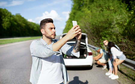 Young guy standing on road next to broken car, looking for mobile network signal, trying to call...
