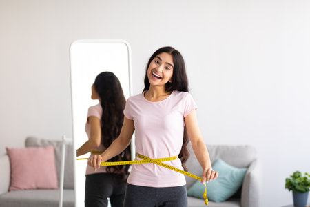 Indian woman measuring her waist with tape in front of mirrorの写真素材