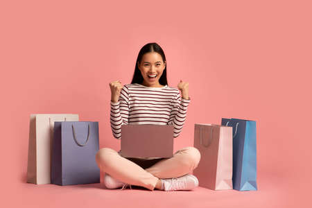 Online Sales. Overjoyed Asian Woman With Laptop Sitting Among Bright Shopping Bagsの写真素材