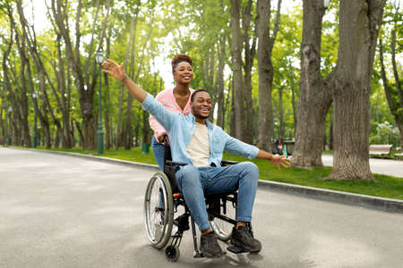 Cheerful young impaired couple with pretty woman and millennial guy in wheelchair going for walk at park togetherの写真素材