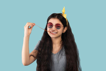 Portrait of playful Indian teen girl in casual wear and sunglasses taking bubble gum out of her...