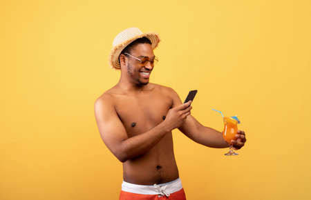 Funky guy with bare torso taking a mobile photo of his tropical cocktail on yellow studio backgroundの写真素材