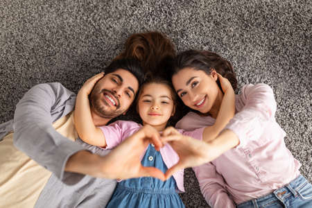 Happy loving family. Arab parents and liitle daughter lying on floor carpet and holding hands in heart shape signの写真素材