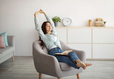 Gorgeous Indian woman stretching in comfy armchair while reading indoorsの写真素材