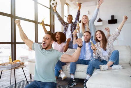 Group of young multiracial friends watching soccer game on TV with snacks and beer, cheering for favorite team at homeの写真素材