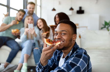 Handsome black guy eating slice of pizza, group of his multiethnic friends with smartphone on backgroundの写真素材