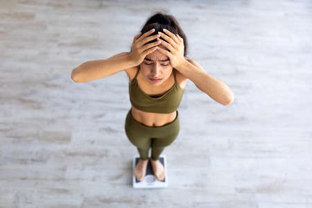 Upset young Indian woman unhappy about her weight, standing on scales, holding head in disappointment, above viewの写真素材