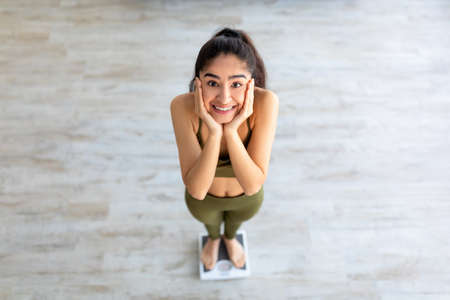 Pretty Indian woman on scales holding her face in excitement, weighing after successful slimming at home, above viewの写真素材