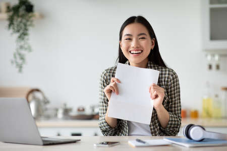 Cheerful asian woman showing empty paper, kitchen interiorの写真素材