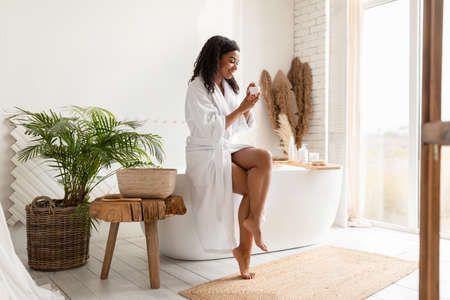 African Woman Holding Moisturizer Cream Doing Skincare Routine In Bathroomの写真素材