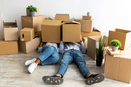 Moving day. Tired african american couple with cardboard boxes on heads, being tired in unpacking thingsの写真素材