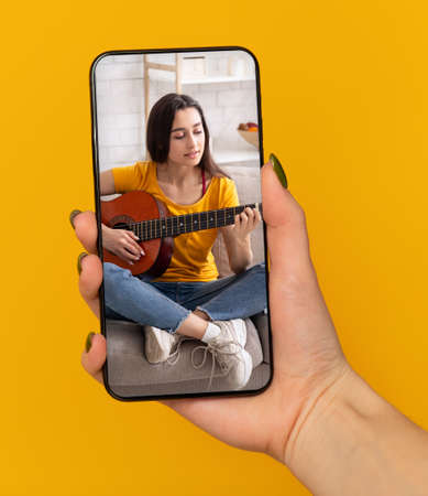 Female hand holding cellphone with online music lesson on screen, learning how to play guitar on web, orange backgroundの写真素材
