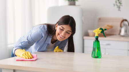 Cheerful young housewife cleaning dining table at kitchen
