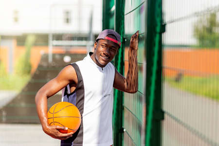 Portrait of millennial African American sportsman with basketball standing near fence at outdoor stadium, copy spaceの写真素材