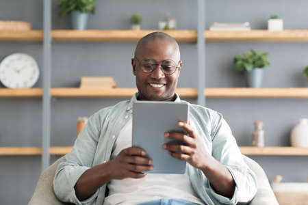 Happy black mature man using digital tablet, sitting in armchair over shelves with accessories,...