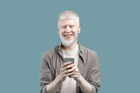 People and technology concept. Excited albino man using smartphone, standing isolated over blue studio backgroundの写真素材