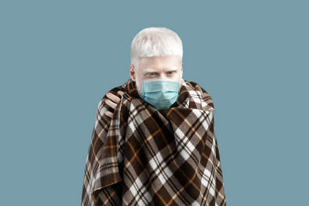 Albino man with pale skin and white hair wearing protective mask and wrapped in blanket, feeling unwell,の写真素材