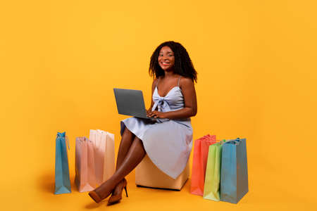 Online shopping. Young black woman using laptop, ordering clothes via internet, surrounded by colorful paper bagsの写真素材