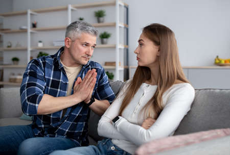 Offended middle-aged woman sitting on sofa after family conflict, her spouse begging for forgiveness at homeの写真素材