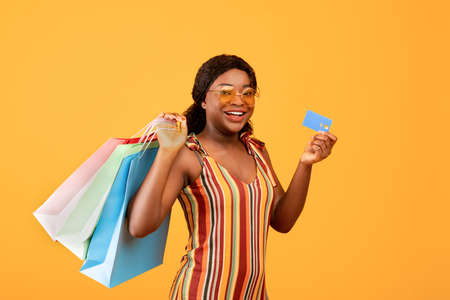 Joyful black woman holding credit card and shopping bags, smiling at camera, choosing contactless...