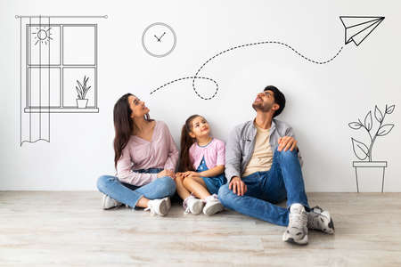 Young family of three imagining interior of their new flat, sitting on floor near white wall with doodle drawingsの写真素材