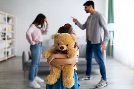 Children and family conflicts concept. Little girl cuddling teddy bear standing against arguing parentsの写真素材