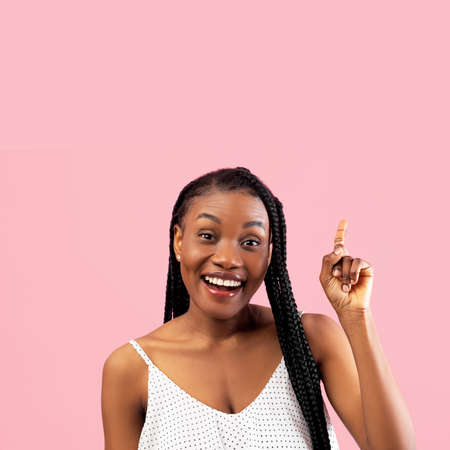 Smiling millennial black woman in white summer dress pointing upwards at empty space on pink studio...
