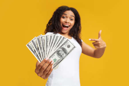 Giveaway concept. Excited young black woman holding a lot of dollar cash, showing big win over yellow studio backgroundの写真素材