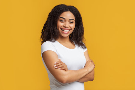 Human emotions. Happy young african american woman with curly hair posing with crossed arms over yellow backgroundの写真素材
