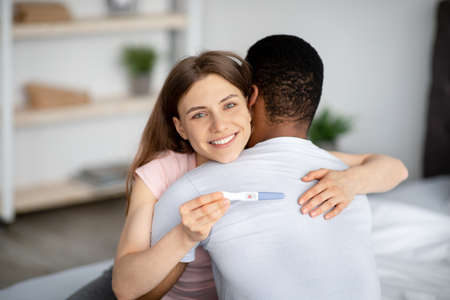 Married multiracial couple happy with positive result of pregnancy test, hugging each other at homeの写真素材