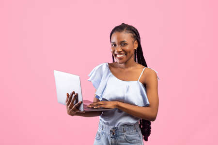 Young African American woman with afro bunches using laptop computer, working or studying online on pink backgroundの写真素材