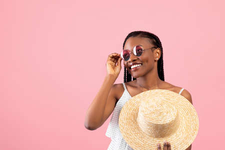 Pretty black lady with afro bunches wearing trendy summer outfit, holding straw hat, looking aside...