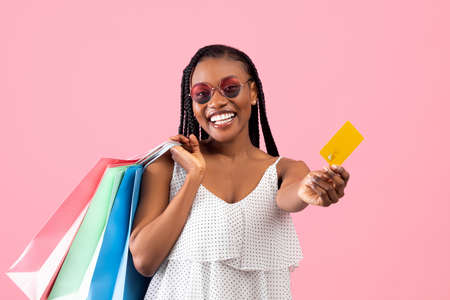 Happy customer. Joyful black lady in summer wear holding colorful shopping bags and credit card over...