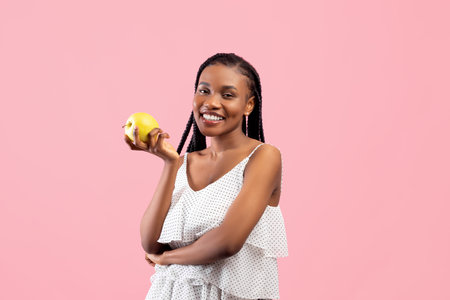 Vitamins for healthy teeth and beautiful skin. Lovely African American woman holding ripe green apple on pink backgroundの写真素材