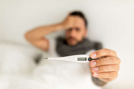 Sick young man showing thermometer to camera, lying on bed and touching forehead. Selective focus, top viewの写真素材