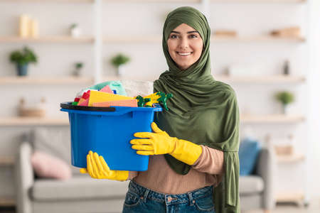 Cheerful young islamic housewife holding bucket with cleaning supplies toolsの写真素材