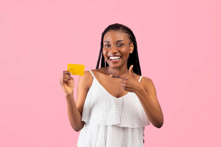 Time for shopping. Happy black lady pointing at credit card and smiling at camera, planning what to buy, pink backgroundの写真素材